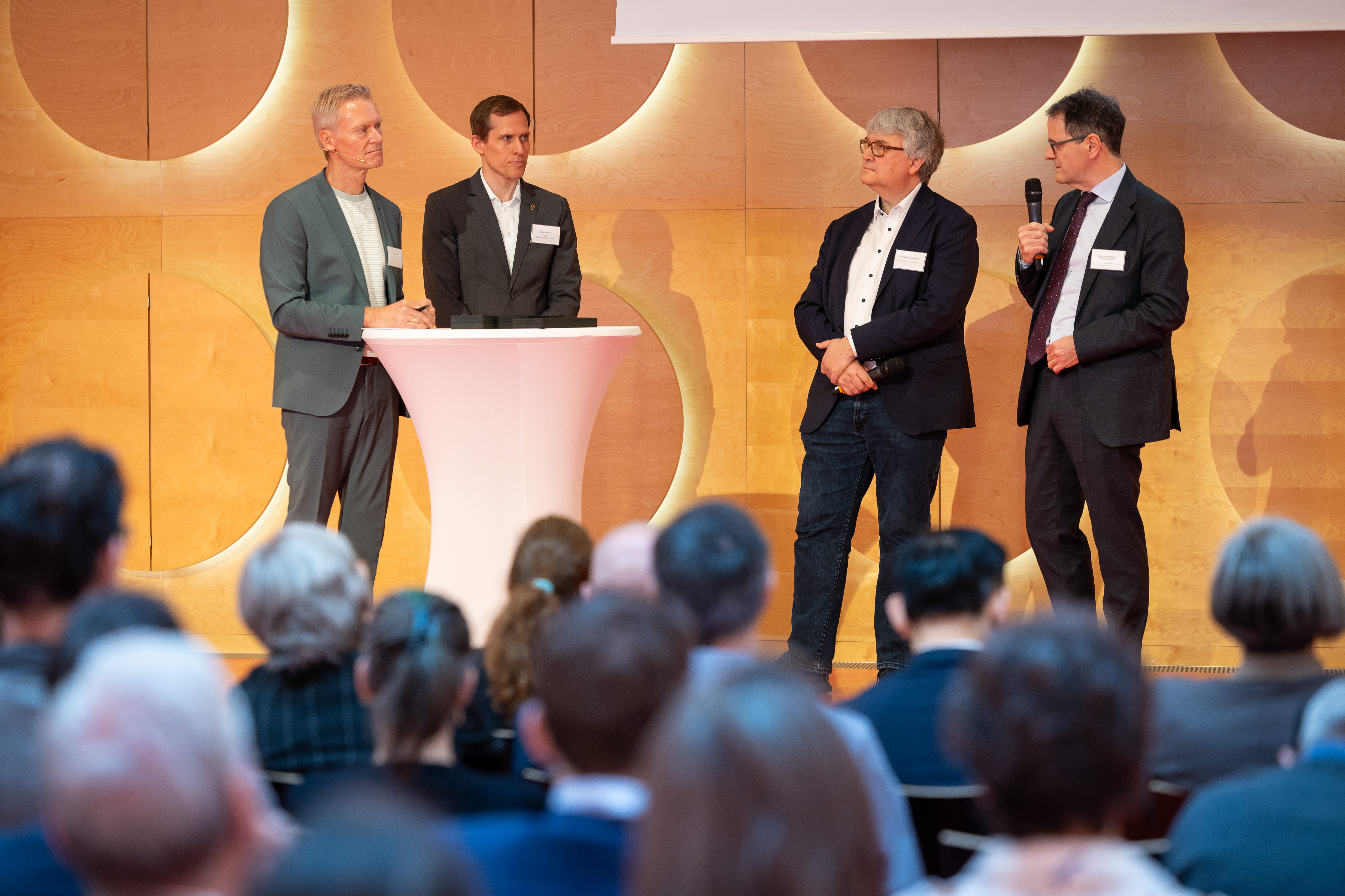 Vier Männer im Anzug stehen um einen Stehtisch vor einem holzvertäfelten Hintergrund. Einer von ihnen, Ministerialdirektor Michael Kleiner, spricht in ein Mikrofon.