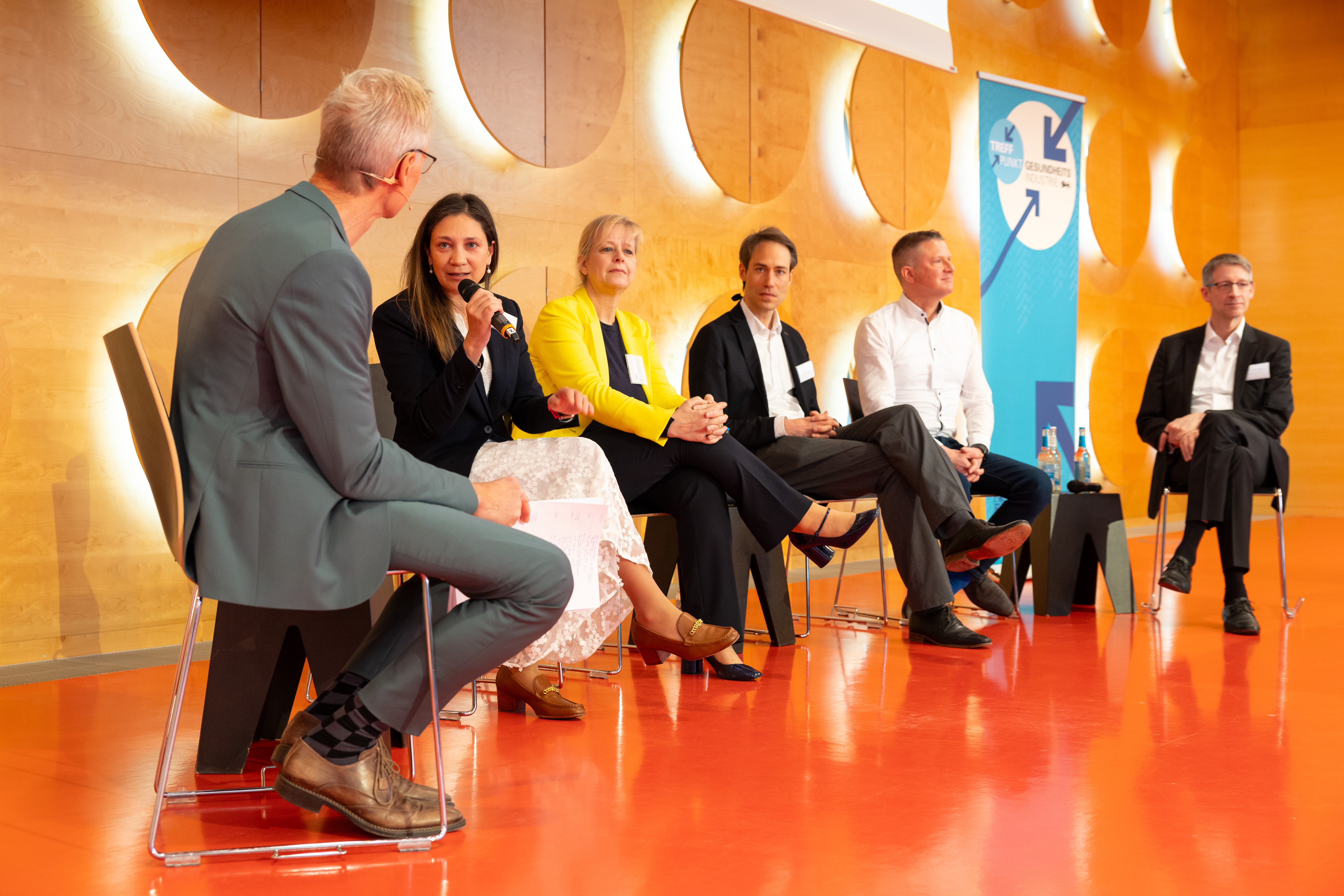 Sechs Personen sitzen auf einer Bühne und diskutieren. Eine Frau spricht in ein Mikrofon.