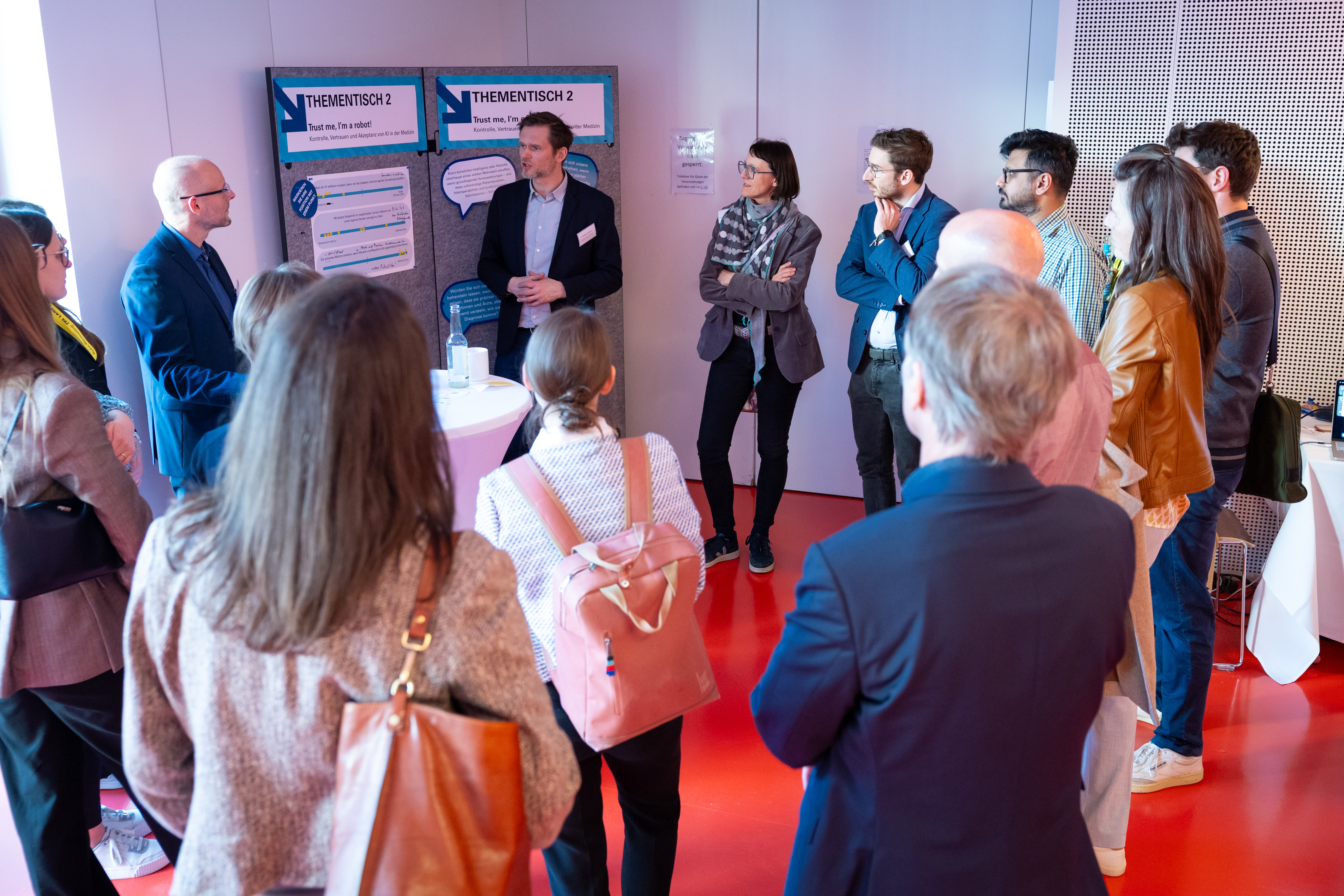 Eine große Personengruppe steht um einen Thementisch herum und diskutiert. Im Hintergrund Pinnwände mit Postern.