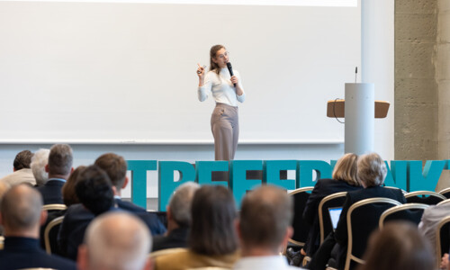 Science Slam auf der Bühne beim Treffpunkt Gesundheitsindustrie 2025 Eine Frau hält einen Vortrag auf der Bühne vor Publikum und spricht mit Mikrofon, im Hintergrund große Buchstaben mit dem Wort „TREFFPUNKT“.