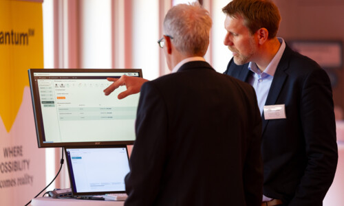 Zwei Männer von hinten gesehen unterhalten sich vor einem Bildschirm. Einer der beiden zeigt auf diesen.