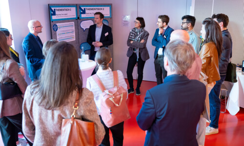 Eine große Personengruppe steht um einen Thementisch herum und diskutiert. Im Hintergrund Pinnwände mit Postern.