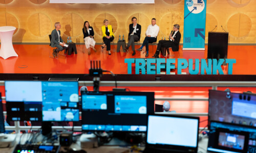 Blick aus einer erhöhten Position auf die Bühne. Im Vordergrund mehrere unscharfe Bildschirme, im Fokus die Bühne mit dem Moderator und fünf Referent:Innen.