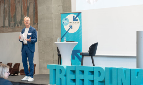 Moderator auf der Bühne beim Treffpunkt Gesundheitsindustrie 2025 Ein Mann steht auf einer Bühne neben einem Stehtisch und einem Banner mit der Aufschrift „Treffpunkt Gesundheitsindustrie“. Vor der Bühne große Buchstaben mit dem Wort „TREFFPUNKT“.