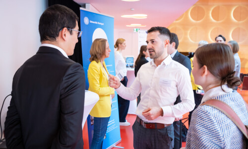 Eine Frau und zwei Männer unterhalten sich. Im Hintergrund sind weitere Gespräche zu sehen.
