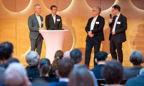  Vier Männer im Anzug stehen um einen Stehtisch vor einem holzvertäfelten Hintergrund. Einer von ihnen, Ministerialdirektor Michael Kleiner, spricht in ein Mikrofon.