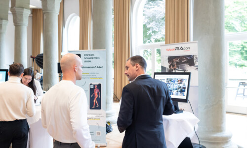 Austausch an Ausstellungsständen beim Treffpunkt Gesundheitsindustrie 2025 Zwei Männer stehen an einem Messestand und unterhalten sich vor Informationsplakaten und einem Bildschirm. Im Hintergrund weitere Teilnehmende und Aussteller.