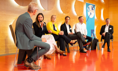 Sechs Personen sitzen auf einer Bühne und diskutieren. Eine Frau spricht in ein Mikrofon.